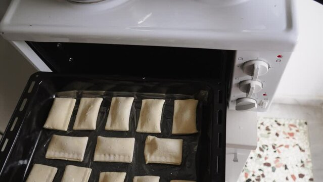 Unrecognizable woman puts homemade raw semifinished baked goods on baking sheet into compact oven. Housewife prepares delicious rectangles filled with puff pastry in tabletop stove at home.