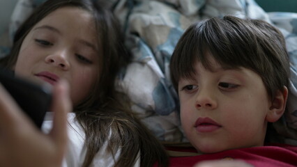 Obraz premium Children staring at phone screen lying in bed, close-up faces of brother and sister watching content online, absorbed by entertainment media