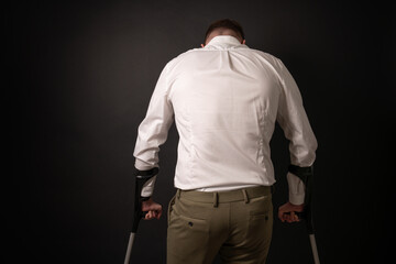Man in white shirt looking down being sad while using crutches, seen from behind.