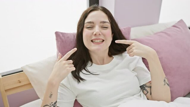 Cheerful young woman in pyjamas pointing at her healthy teeth, sitting in bedroom. dental health in good vibes!