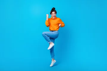 Fototapete Full length photo of cute lucky woman dressed orange sweater rising fists jumping high isolated blue color background © deagreez