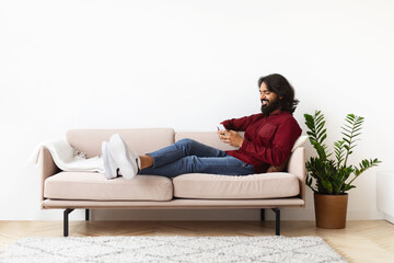 Cool millennial indian guy reclining on couch with smartphone