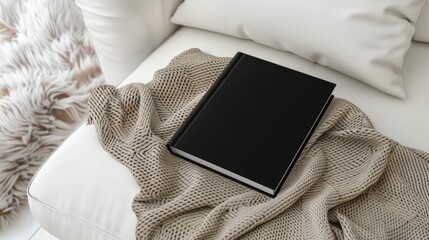 a closed slim B-format black softcover book placed prominently on a white chic sofa, accompanied by an oatmeal-colored wool blanket, creating a cozy and inviting atmosphere.