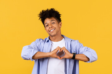 Happy black guy making heart sign with hands on yellow background - obrazy, fototapety, plakaty