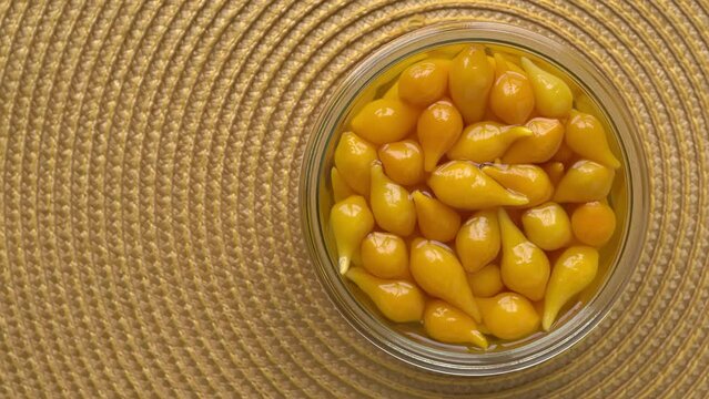 Top view of pickled whole Biquinho Yellow chili in a glass jar on a yellow background. Sour and sweet Asian-style pickled homegrown chilies. Table spin.