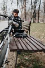 Stylish male entrepreneur engaged in remote work with a bicycle nearby in an urban park setting, focusing attentively on his laptop.