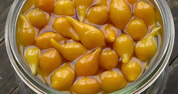 Close up view of pickled whole Biquinho Yellow chili in a glass jar on a rustic wooden table. Sour and sweet Asian-style pickled homegrown chilies. Table spin.