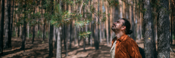 Portrait of a young man with a backpack on a walk in a pine forest on a sunny day. The guy breathes fresh air in a coniferous forest © Anna