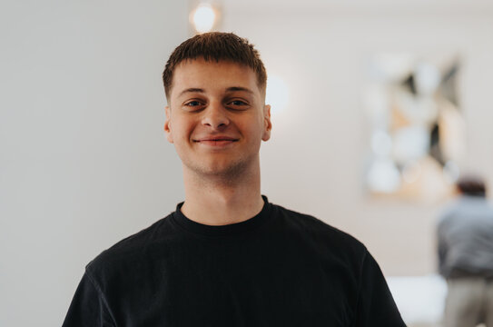 A young adult male with a friendly smile, wearing a casual black t-shirt, poses for a portrait with a softly blurred interior background.