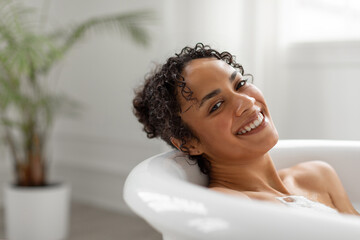 Relaxed black woman enjoying foamy hot bath, leaning on tub and smiling at camera, relaxing at...