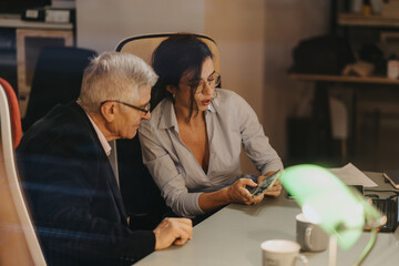 Two professional senior colleagues collaborate on a project, focusing intently on a smart phone in...