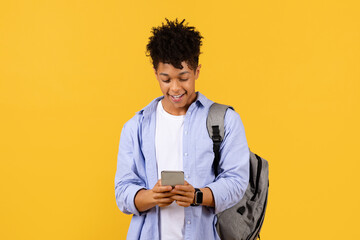 Happy black male student with smartphone and backpack against yellow background