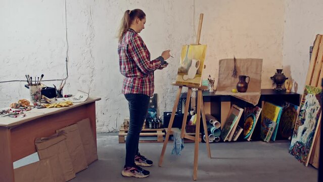 Painter does a still life painting with a thin brush. Young femail artist studio. Natural lighting from a big window.  