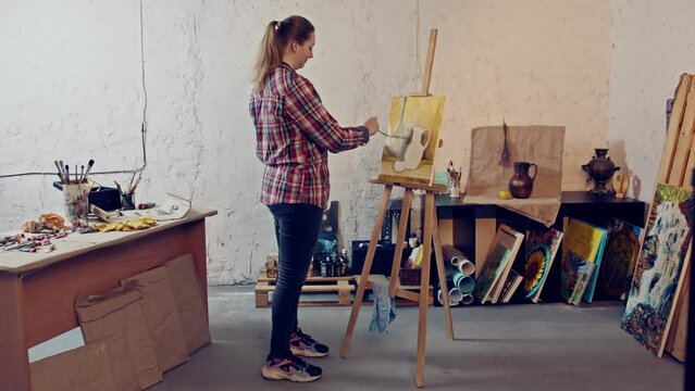 Woman artist is looking at the canvas while painting. Warm colors still life painting in a workshop. 