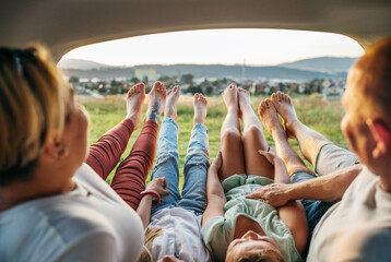 Four-person family lying in the car trunk and fooling around raising feet up. The young caucasian couple has an auto trip break. Family values, traveling, and friendship concept. - obrazy, fototapety, plakaty