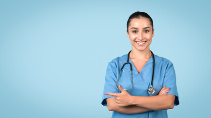 Happy nurse pointing left at free space, wearing blue scrubs - obrazy, fototapety, plakaty