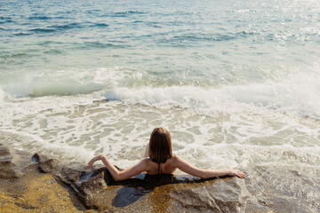Fototapeta na wymiar A woman lies on a rock, arms spread wide, as gentle waves caress the shore