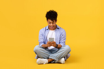 Joyful black guy using smartphone while sitting cross-legged on yellow background