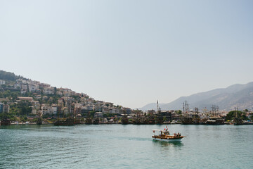 A small boat sails across calm waters with a bustling hillside town and ships in the background