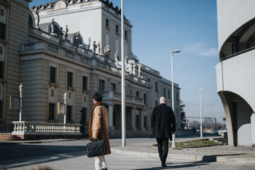 Two businesspeople in formal attire walking outside on a sunny day, juxtaposing classic architecture with modern design.