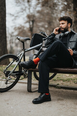 Naklejka premium Handsome businessman working remotely while sitting on a park bench with his bicycle, enjoying a natural urban setting.