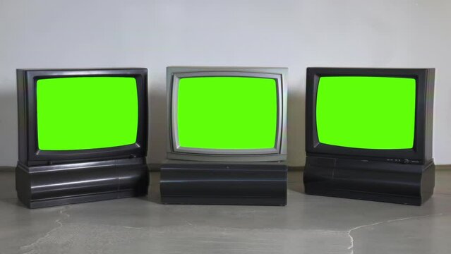 Three classic televisions with green screens side by side on a grey floor against a white wall.