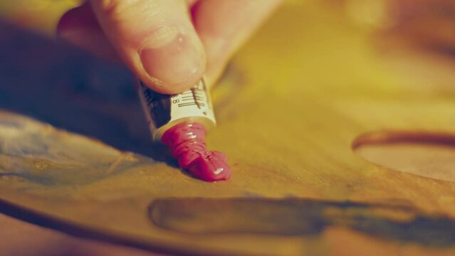 Artist squeezing pink paint from a tube onto a palette. Artist’s wooden palette with different colors. 