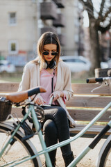 Fashionable young woman checking her smart phone while sitting on a bench in a sunny park, with her bicycle beside her.