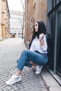The girl sat down next to the wall in stylish clothes. Girl posing on the background of a wall in the city
