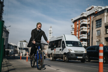 Focused male city commuter riding a bicycle on an urban road with vehicles in the background during...