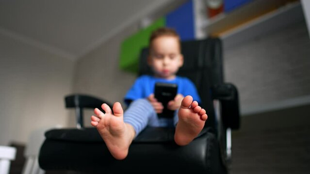 Bare little feet of a baby boy sitting in a big office chair. Close up. Blurred image of a child playing with phone. Low angle view.