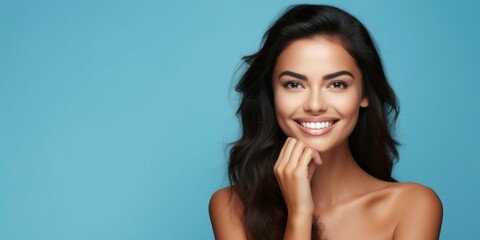 Radiant young brunette woman smiling, touching face, on blue background. Concept of skincare beauty, beauty, joyful demeanor, and positive attitude. Wide banner with copy space - obrazy, fototapety, plakaty