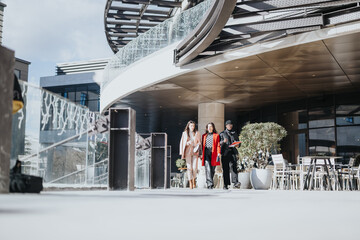 Two women and a man in stylish autumn attire stroll through a contemporary, open-air plaza with modern buildings and outdoor seating.