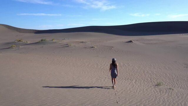 A young Latina woman in a dress and cap walking backwards through a huge dune field in Patagonia.