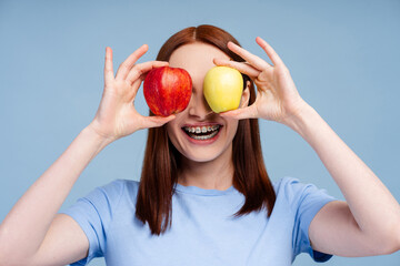 Ginger smiling woman with braces in blue shirt covering eyes with two apples isolated