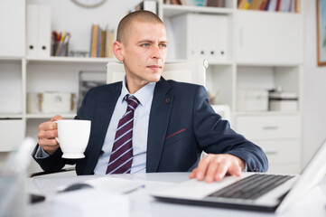 Young businessman working on laptop while sitting by desk in white modern office