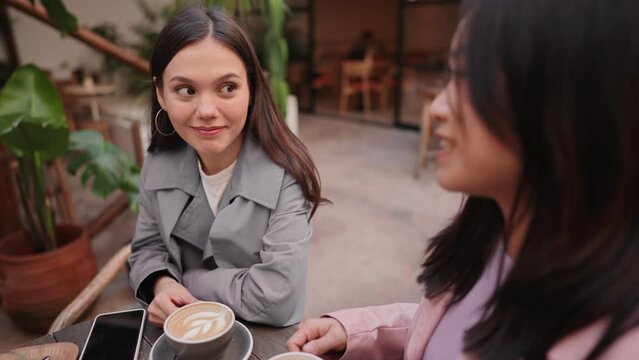 Two young women share a moment over coffee at a cafe, holding a drink while listening intently to her friend.