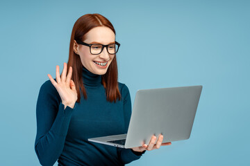 Red haired woman with braces wearing eyeglasses holding laptop computer, waving hand