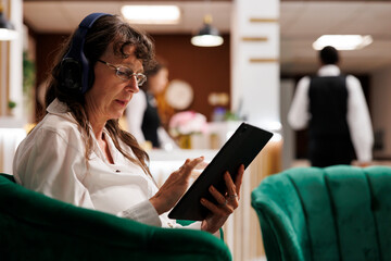 Detailed shot of senior woman relaxing with headphones and surfing the net on tablet in hotel...