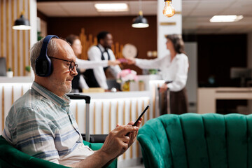 Detailed view of elderly male guest relaxing in cozy lounge area with tablet and wireless headset....