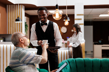 African american employee serving coffee to eldery male tourist with tablet in luxury hotel...
