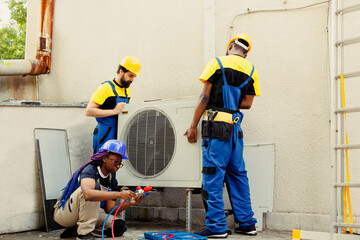 Diverse team of seasoned wiremen installing new external air conditioner for customer. Licensed servicemen tasked to optimize new HVAC system's performance, ensuring it runs at perfect efficiency
