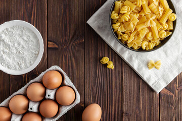 Different kinds of raw pasta with eggs on wooden background.
