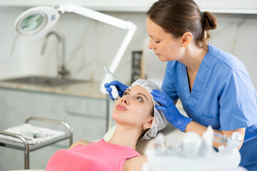 Adult woman cosmetologist performs facial microcurrent procedure with machine to young female client