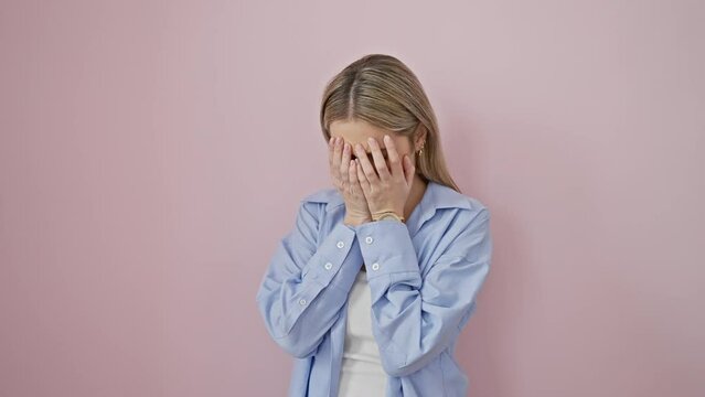 Depressed young blonde woman covering tear-streaked face with hands, crying over isolated pink background