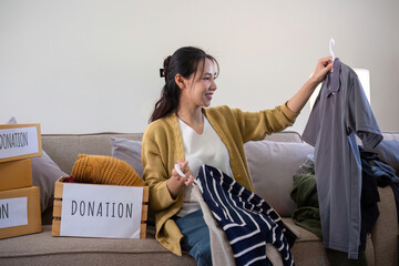 Cute Asian woman sits on a sofa sorting unwanted clothes to donate.
