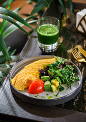 Omelette with fresh salad, arugula, avocado and tomato on plate on table with green smoothie....
