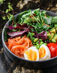 Buddha bowl salad with salmon fish, arugula, spinach, avocado, tomatoes, green edamame beans, eggs on marble background. Healthy salad, dieting, top view, close up
