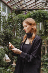 Elegant caucasian woman in black evening jumpsuit uses a stylus with a tablet in a lush garden....