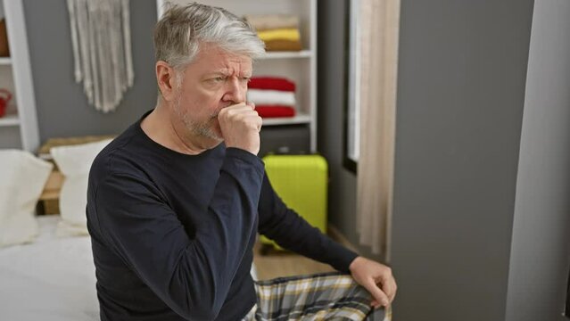 Mature grey-haired man coughing while sitting on a bed in a bedroom, presenting illness symptoms indoors.